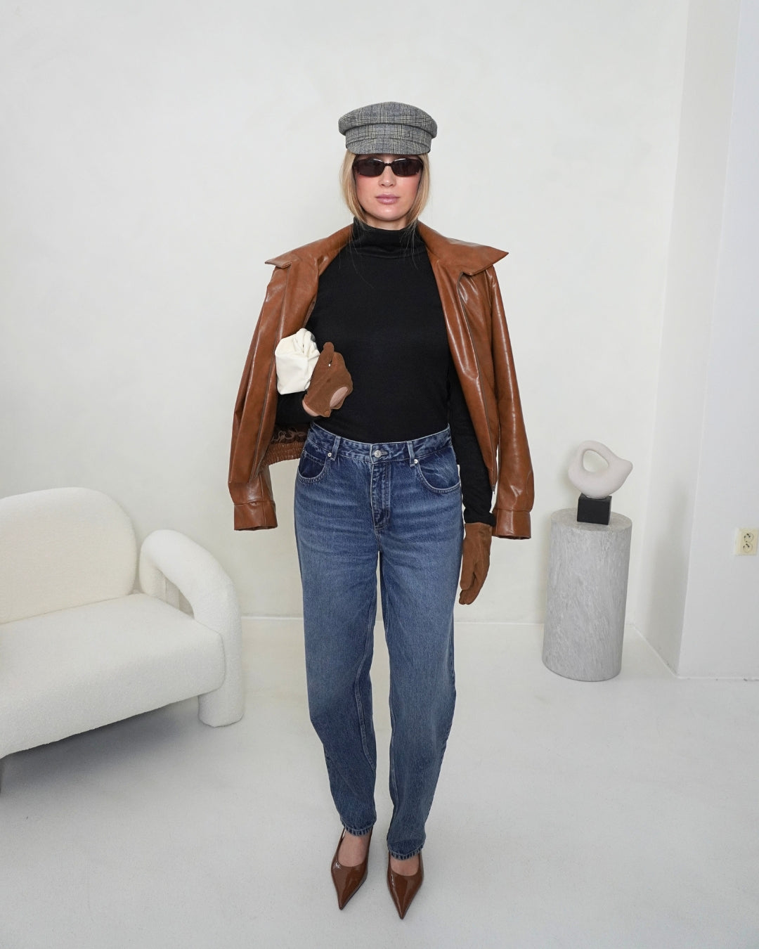 Black SELENE Turtleneck by THÉFI layered under the brown HERA Faux Leather Jacket, styled with blue jeans and a grey cap.
